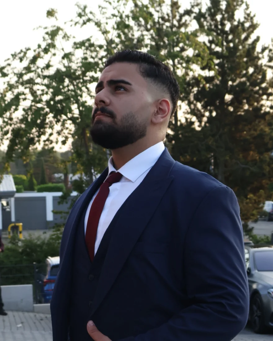 Umut Yagmur im dunkelblauen Anzug mit roter Krawatte, draußen vor Bäumen